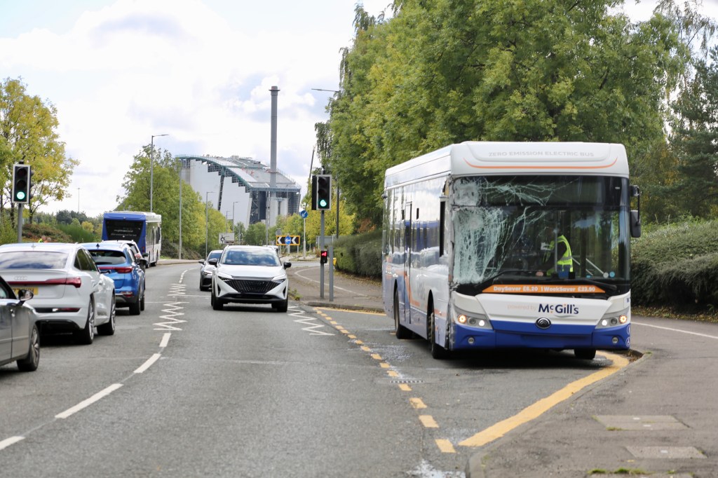 A damaged electric bus operated by McGill's, stopped on a road with traffic lights and vehicles in the background, with a power station visible in the distance.