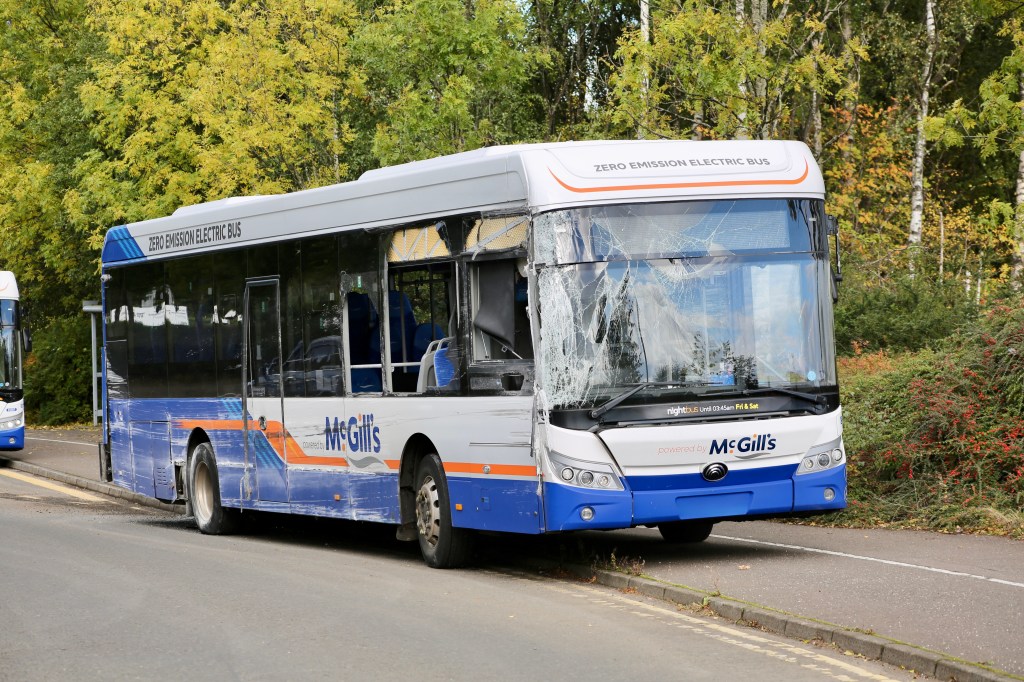 A damaged McGill's zero emission electric bus parked by the roadside, with a cracked windscreen, surrounded by trees and greenery.