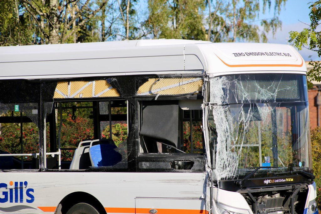 A front view of a damaged electric bus with a cracked windshield, parked in a green area.