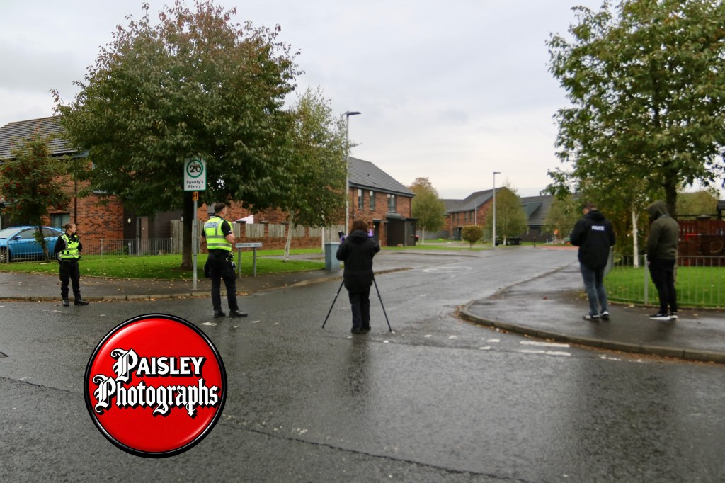 Police officers and bystanders gather at a residential street corner, with a 20 mph speed limit sign visible and houses in the background.