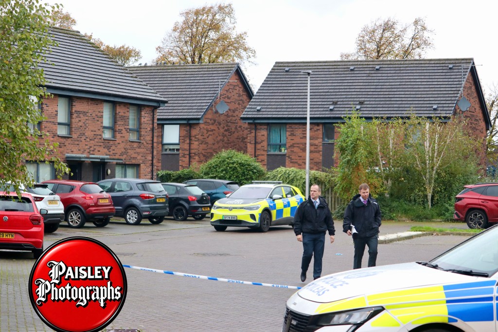 Police officers walking past a police cordon at a residential area, with parked cars and houses in the background.