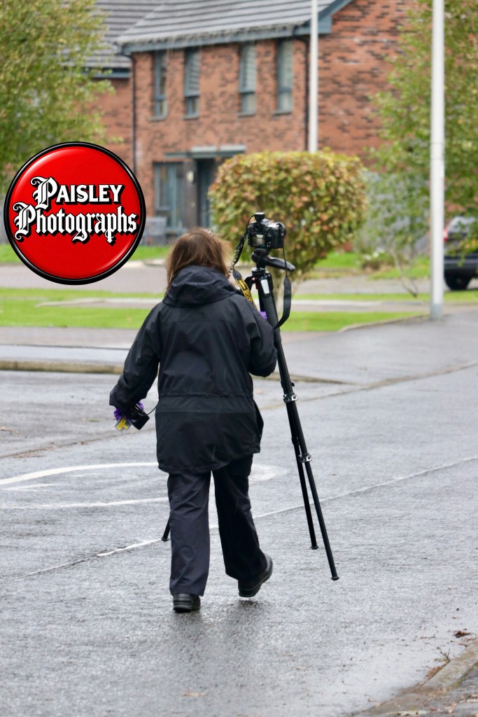 A person in a black coat walks along a wet road, carrying a camera on a tripod and holding some items in their hand, with residential houses in the background.
