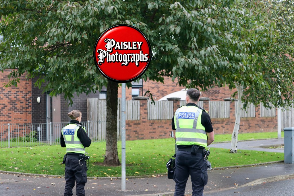 Two police officers standing near a tree and a sign for 'Paisley Photographs' in a residential area.