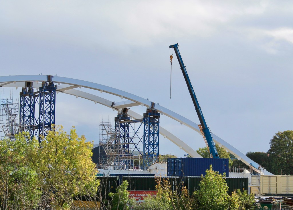 Construction site featuring a large steel arch structure with a crane and scaffolding in the foreground.
