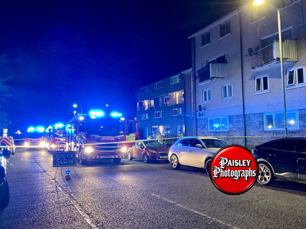 Emergency vehicles with flashing blue lights parked at night along a residential street, with a nearby police sign warning to slow down.