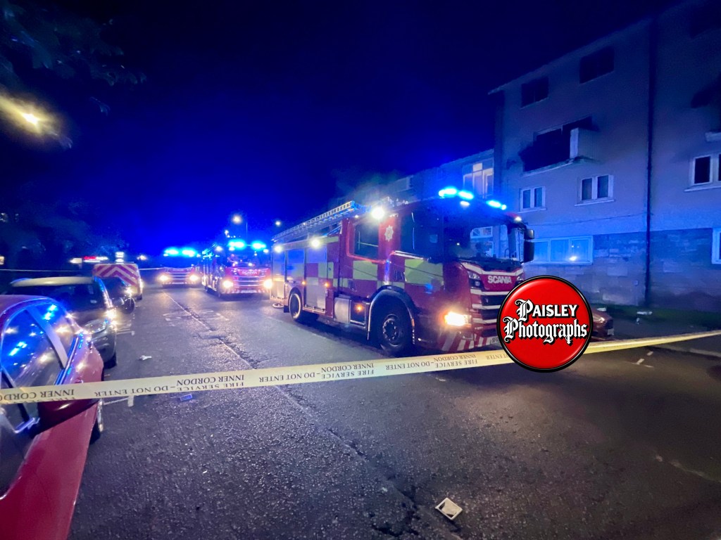 Fire engine parked on a street at night, surrounded by police vehicles and illuminated by blue lights, with a 'Fire Service Do Not Enter' tape across the scene.
