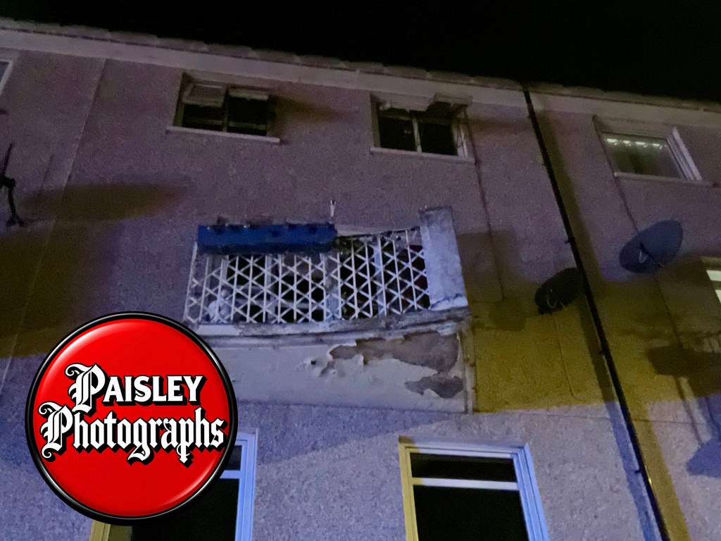 A close-up view of a damaged upper exterior wall of a building at night, featuring a deteriorating balcony with a blue panel and two windows, along with satellite dishes.