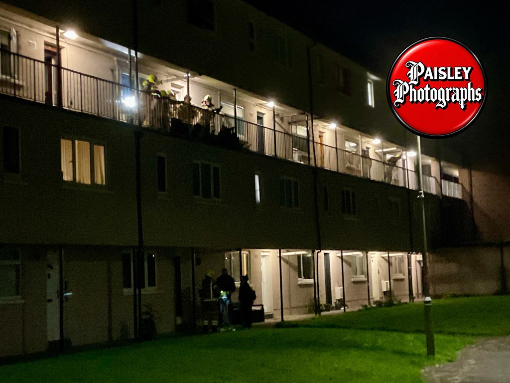 A night view of a residential building with people on the balcony and activity occurring outside.